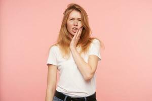 Studio shot of young redhead female with natural makeup keeping palm on her face and squinting her eyes while looking at camera, isolated over pink background photo