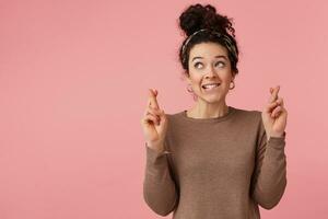 Portrait of a young girl with dark curly hair, biting her lip, fingers crossed, hopes that she will be lucky on the exam. Looking up isolated over pink background. photo