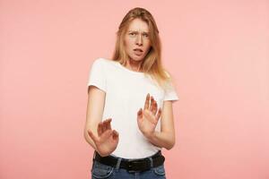 Studio photo of young confused redhead woman with casual hairstyle raising palms in protective gesture while looking scaredly at camera, isolated over pink background