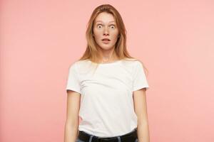 Studio photo of young amazed long haired redhead lady rounding surprisedly her green eyes while looking at camera, isolated over pink background in white basic t-shirt