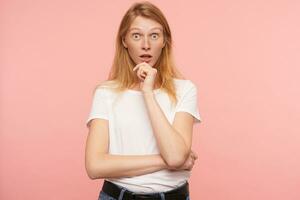 Studio photo of young bemused redhead female holding her chin with raised hand while looking surprisedly at camera with wide eyes opened, isolated over pink background