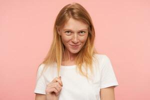 Portrait of lovely young shrinking female in white t-shirt pulling her foxy hair while looking positively at cmera with gentle smile, standing over pink background photo