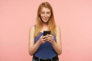 Studio photo of young attractive female with loose foxy hair holding smartphone in hands and looking wonderiongly on screen, standing against pink background