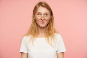 Portrait of charming young redhead lady with natural makeup looking positively at camera and smiling pleasantly, posing over pink background with hands down photo