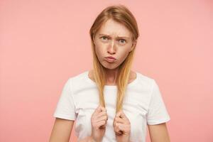 Funny shot of young green-eyed long haired redhead female dressed in white basic t-shirt pursing her lips and grimacing face while fooling over pink background photo