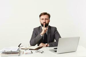 Indoor photo of serious young bearded businessman working at office with his laptop and notebook, sitting at table over white background and raising hand in hush gesture