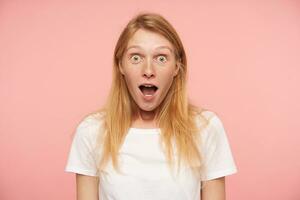 Bemused young lovely redhead woman with casual hairstyle looking surprisedly at camera with wide eyes and mouth opened, isolated over pink background photo