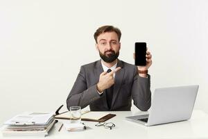 bearded businessman, top manager sitting at desktop in office, looking at camera with sullen facial expression, dressed in an expensive suit with a tie, pointing with finger to his device. photo