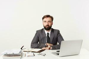 Studio shot of annoyed young brunette male with beard sitting at working table and keeping folded hands on countertop, looking upwards with pout while posing over white background photo