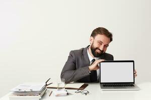 Cheerful young handsome bearded businessman with short brown hair wearing grey suit while sitting at working table over white background, showing screen of his laptop and smiling broadly photo