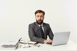 Shocked attractive bearded businessman, top manager sitting at desktop in office, looking at camera with wide open mouth, heard the shocking news. Dressed in an expensive suit with a tie. photo