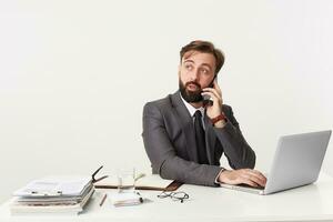 retrato de un joven atractivo barbado empresario que se discute un importante negocio problema en el teléfono. sentado a escritorio en oficina, trabajando para su computadora portátil, vestido en un traje con atar. foto