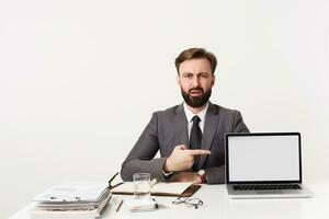 Confused young handsome brunette male with beard and short haircut posing over white background at working table and showing with forefinger on his laptop photo