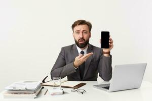 Portrait of shocked attractive bearded businessman, manager sitting at desktop in office, looking at camera in disbelief, dressed in an expensive suit with a tie, pointing to his smartphone. photo