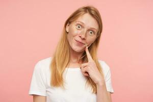 Indoor shot of positive young pretty redhead female with natural makeup rounding her green eyes and holding forefinger on cheek while posing over pink background photo