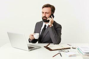 Serious bearded brunette man in formal clothes keeping white cup in raised hand while having phone conversation, frowning eyebrows while looking at screen of his laptop, isolated over white background photo