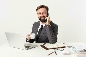 Studio shot of pretty positive bearded brunette male with short haircut sitting at working table with cup of coffee, making call with his smartphone and looking aside with broad smile photo