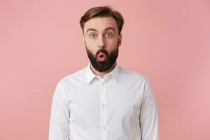 Photo of dazed handsome bearded man, didn't expect to hear the shocking news, wearing a white shirt. Looking at the camera with wide open mouth isolated over pink background.