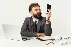 Displeased young brunette man with beard wearing trendy hairstyle and formal clothes while working at office with notebook and laptop, looking with pout on smartphone in his raised hand photo