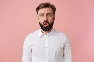 Photo of frown attractive dazed bearded man, wearing a white shirt. Looking at the camera with wide open mouth isolated over pink background.