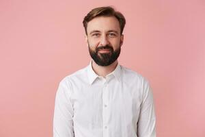 Portrait of young calm smiling handsome bearded man, wearing a white shirt. Looking at the camera isolated over pink background. photo