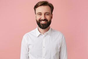 retrato de alegre joven hermoso barbado hombre, vistiendo un blanco camisa. mirando a el cámara y sonriente aislado terminado rosado antecedentes. foto