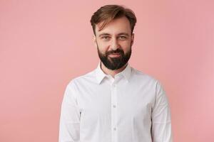 Portrait of young calm handsome bearded man, wearing a white shirt. Looking at the camera and smile wit shy isolated over pink background. photo