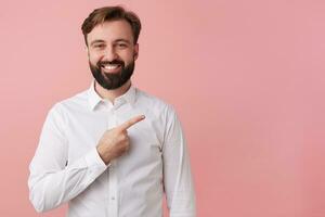 Happy handsome bearded young man, wearing a white shirt. Wants to tell cool news. Broadly smiling, draws your attention pointing to the copy space on the right isolated over pink background. photo