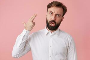 Young bearded man tries to kill himself with a headshot with two fingers isolated over pink background. photo