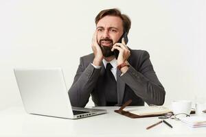 Bemused young brunette man with beard wearing formal clothes while working over white background, leaning head on raised hand and looking confusedly on screen of his laptop photo