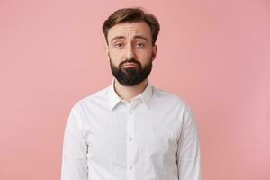 Portrait of a handsome bearded man who is upset about something, wearing a white shirt. Looking at camera with sullen facial expression isolated over oink background. photo