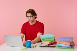 Photo of young frowning student in glasses wears in red t-shirt, man sits by the table and working with laptop and books, isolated over pink background. Looks doubtful and displeased.