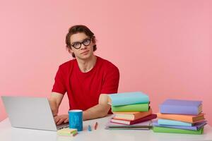 Studio photo of young man in glasses wears in red t-shirt, student sits by the table and working with books and notebook, prepared for exam, having serious look, isolated over pink background.