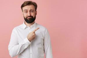 Frowning unhappy man wants to draw your attention pointing with finger on the copy space on the right, isolated over pink background. photo