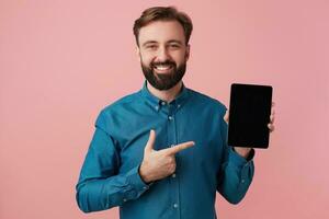 Happy smiling bearded man wants to draw your attention, pointing with finger to his device. Looking at camera in surprise isolated over pink background. photo