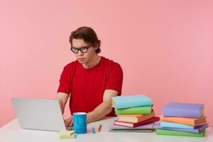 Frowning young student in glasses wears in red t-shirt, man sits by the table and working with laptop and books, looks at the monitor with surpraised, something wrong, isolated over pink background. photo