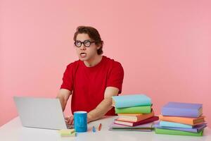 Irritated young student in glasses wears in red t-shirt, man sits by the table and working with laptop and books, very tired and angry, hate exams, loks up isolated over pink background. photo