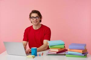 Portrait of smiling man student in glasses wears in red t-shirt, sits by the table and working with notebook and books, prepared for exam, broadly smiles isolated over pink background. photo