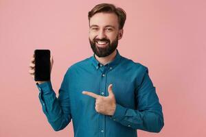 Happy smiling bearded man wants to draw your attention, pointing with finger to his smarthpone, wearing a denim shirt. Looking at camera in surprise isolated over pink background. photo
