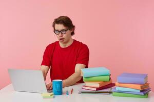 infeliz joven estudiante en lentes usa en rojo camiseta, hombre se sienta por el mesa y trabajando con ordenador portátil y libros, mira a el monitor con sorprendido, alguna cosa equivocado, aislado terminado rosado antecedentes. foto