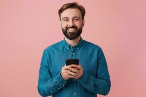 Portrait of a contented attractive bearded young man, wearing a denim shirt, holding a smartphone in his hands, smiling widely and looking at the camera isolated over pink background. photo