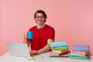 Funny young student in glasses wears in red t-shirt, sits by the table and working with laptop, looks happy and joyful, holding a pencil with his lips and cup in hand, isolated over pink background. photo