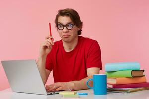 Young student in glasses wears in red t-shirt, man sits by the table and working with laptop, looks at the camera, holds in hand a pencil, have a cool idea, isolated over pink background. photo