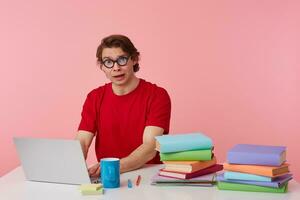 Portrait of sad young student in glasses wears in red t-shirt, sits by the table and working with laptop, looks unhappy, isolated over pink background. photo