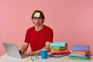 joven cansado hombre en lentes usa en rojo camiseta, con un pegatina en su frente, se sienta por el mesa y trabajando con cuaderno y libros, tristemente mira a el cámara, aislado terminado rosado antecedentes. foto