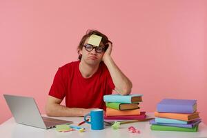 Young tired man in glasses wears in red t-shirt, sits by the table and working with notebook and books, with a sticker on his forehead, sadly looks at the camera, isolated over pink background. photo