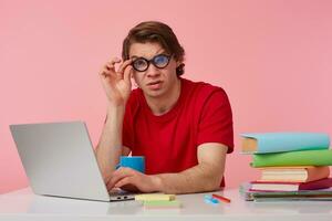 Young student in glasses wears in red t-shirt, man sits by the table and working with laptop, looks at the camera through glasses with disgusted expression, isolated over pink background. photo