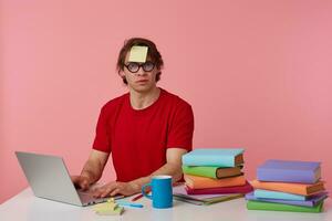 Young man in glasses wears in red t-shirt,with a sticker on his forehead, sits by the table and working with notebook, prepared for exam, having serious look, isolated over pink background. photo