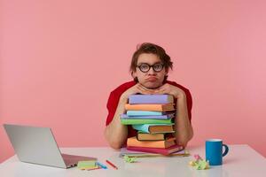Photo of young man in glasses wears in red t-shirt, man sits by the table and working with laptop and books, leaned on a pile of books, isolated over pink background. Looks displeased and unhappy.