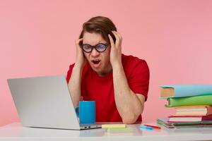 angry young student in glasses wears in red t-shirt, man sits by the table and working with laptop, hols his head and screams, all the work done is not saved, isolated over pink background. photo